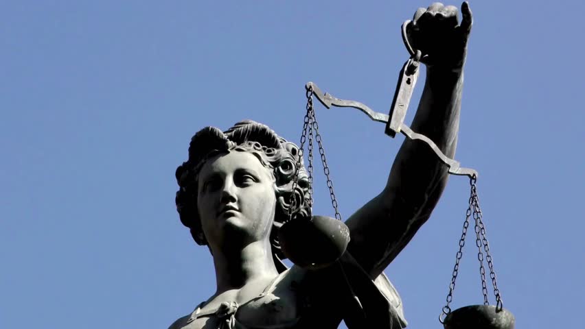 Statue depicting Justice stands tall, holding scales high, surrounded by clear blue sky. Location showcases urban architecture and historical significance during daylight hours.