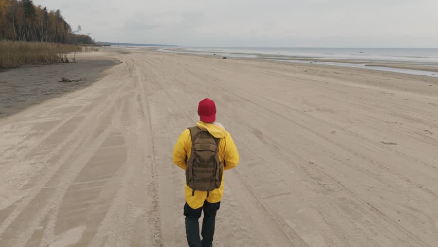 traveler with backpack runs along beach in cloudy weather. drone captures man in yellow raincoat moving along lake.