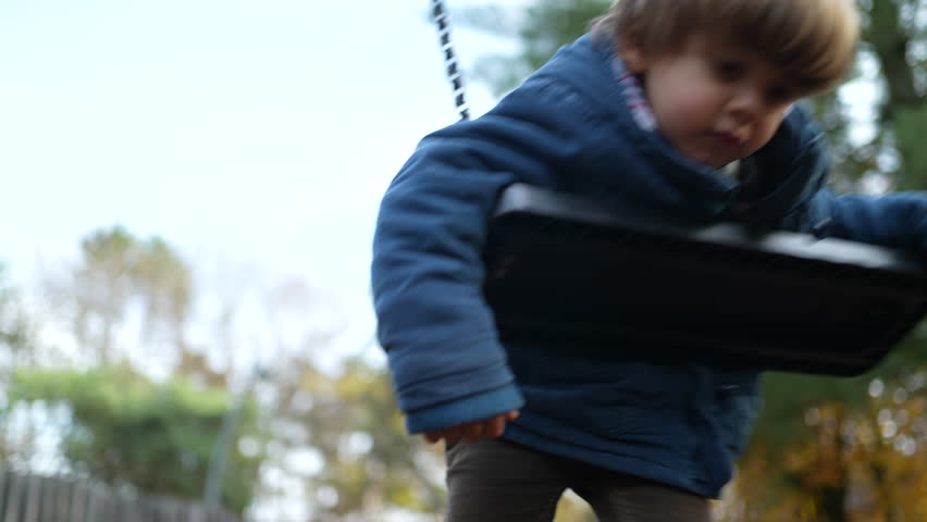 Child leaning on park swing on his stomach outside during autumn fall season day, kid wearing blue jacket