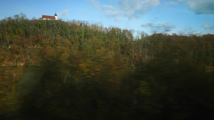Moving view of forested hillside seen from train window with distant building, motion blur, travel perspective, countryside landscape, everyday journey moment
