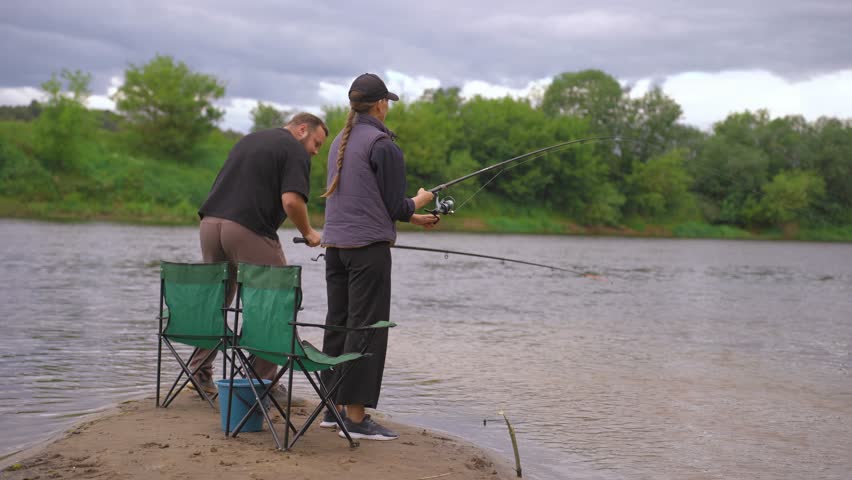 Passionate couple enjoying a quiet day fishing together from the sandy riverbank, with the woman actively reeling in the line while the man relaxes next to her in his chair