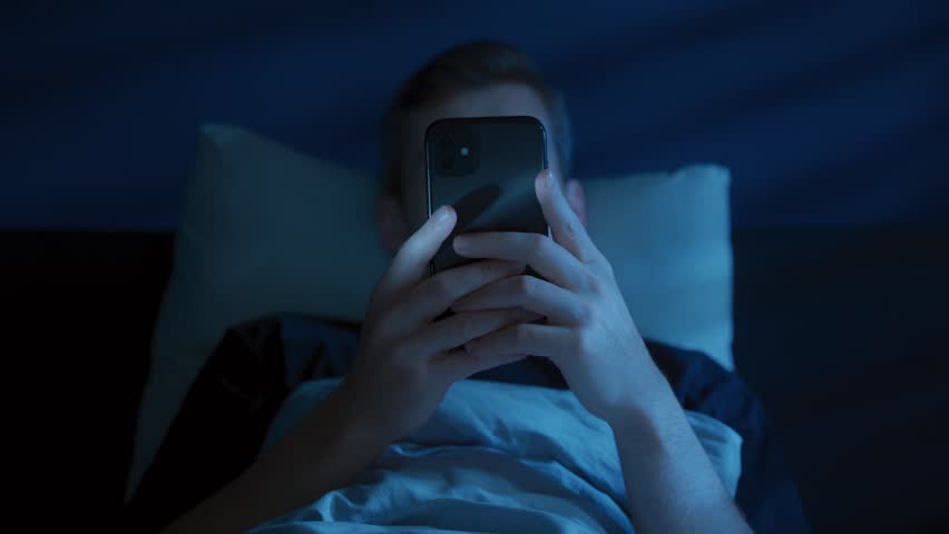 Close up of young man, alone in the dark bedroom, holds his smartphone close to face while scrolling social media and texting late at night. Screen dependence, content consuming or sleep disorder 