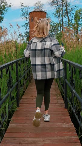 A young woman with blond hair runs casually across a wooden bridge and a stone path towards an old brick tower while enjoying a beautiful sunny day in a picturesque public park vertical video