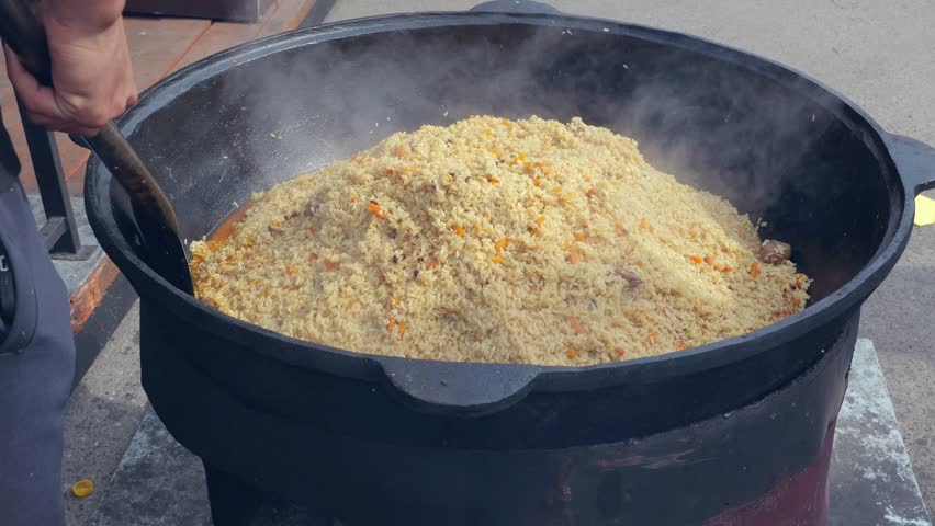 Huge pilaf mound being turned by cook with long ladle, steaming golden rice and carrot flecks in oversized pot, communal catering scene at busy festival, tactile rhythmic