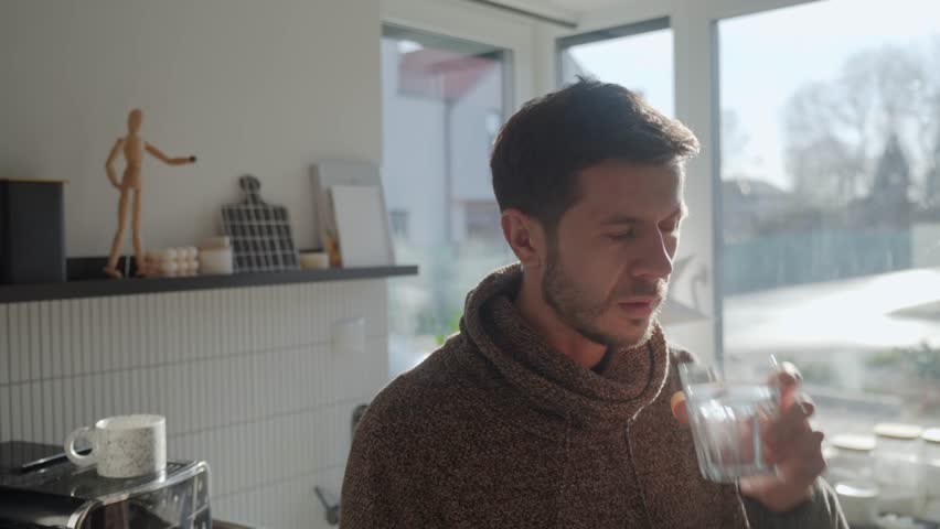 Adult man drinking fresh clean water from glass while standing in kitchen near window with sunlight. Person quenching thirst at home. Concept of hydration and healthy lifestyle