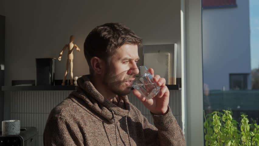 Adult man drinking fresh clean water from glass while standing in kitchen near window with sunlight. Person quenching thirst at home. Concept of hydration and healthy lifestyle