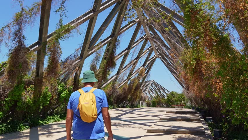 mature man with a backpack exploring a contemporary pedestrian bridge with climbing plants. Sunny day in a public park in santiago, chile