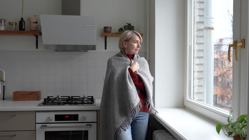 Tired middle aged woman covering in plaid to stay warm in cold unheated apartment, feeling sick coughing and sneezing, seasonal health problems caused by utility issues and low temperature in winter.