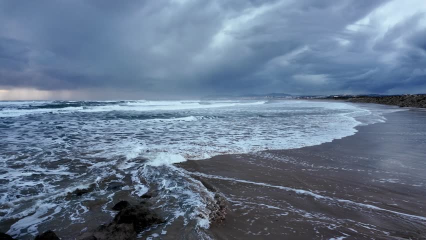 Aerial view of people walking along the curved coastal path in Costa da Caparica on a sunny winter day. Blue ocean, waves, and stone barrier in frame.
