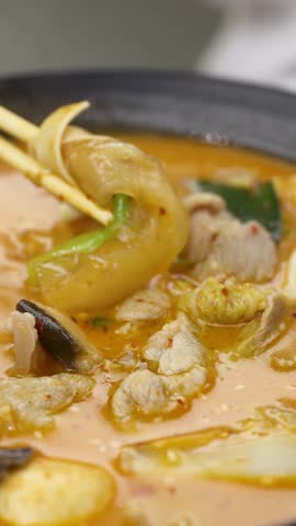 Closeup of chopsticks and spoon lifting vegetables from bubbling spicy hotpot soup, shallow focus