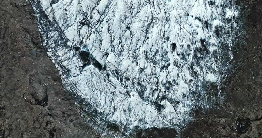 Aerial view of a glacier edge where white ice meets dark volcanic terrain, revealing striking natural patterns, cracks, and contrasting textures.
