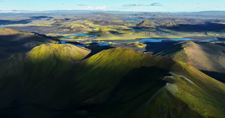 Golden light sweeps across Iceland’s volcanic highlands, revealing moss-covered peaks and bright river channels winding through the vast rugged landscape.