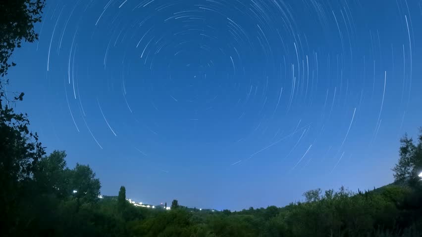 Captivating star trails time-lapse over a serene landscape with a small village and lush green hills under a clear night sky