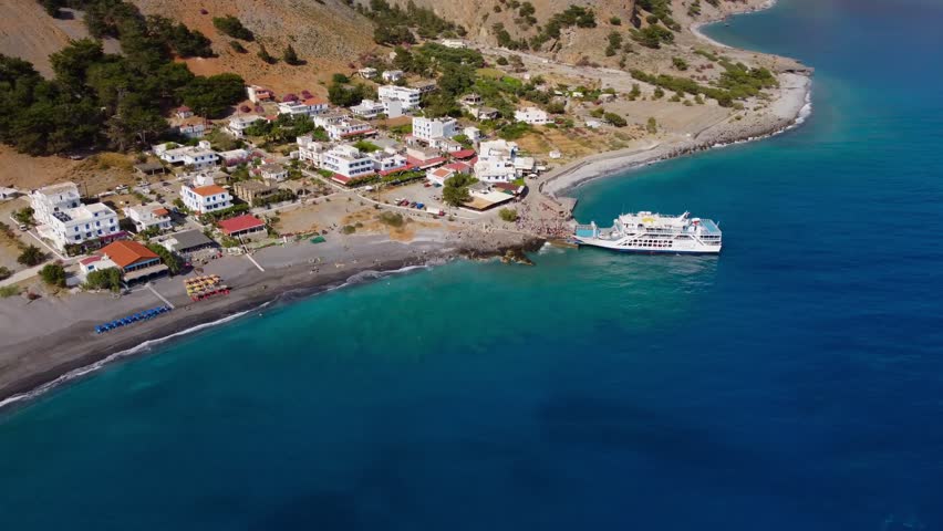 Drone circling coastal village of Agia Roumeli with large ferry. Southern Coast of Crete with beautiful beach and sea views