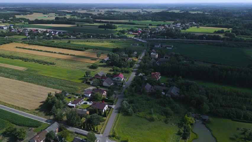 Cinematic drone footage pulling back over a Polish village to reveal expansive, colorful agricultural strips and traditional houses nestled in the lush countryside of the Łowicz region.