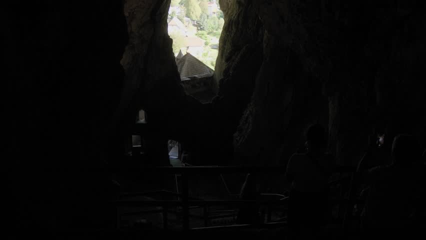 Tilt up from the dark interior of a cave inside Predjama Castle, revealing a bright opening that frames the village and landscape outside.