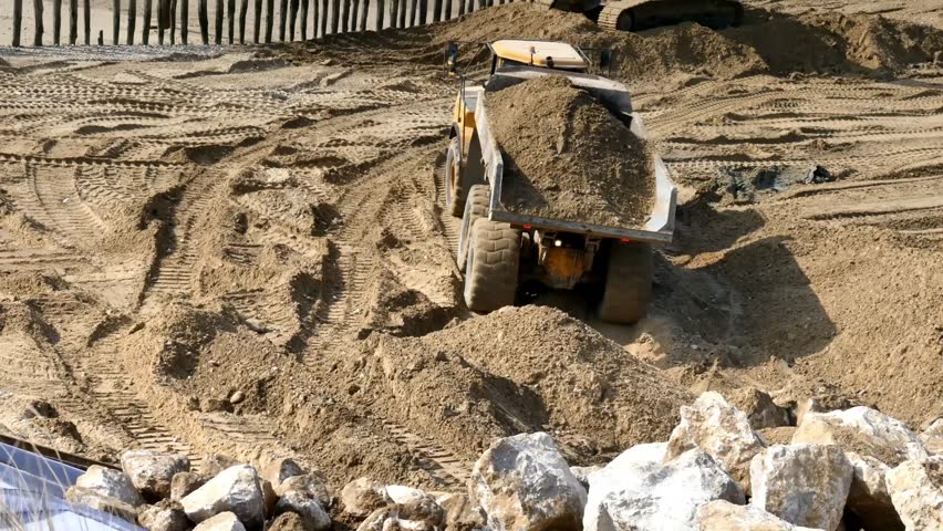 A 4K construction scene showing an excavator bucket dumping a large load of dirt onto a pile at an active earthmoving site.