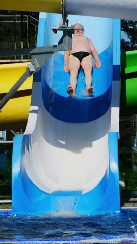 Elderly man descends high slide and joyfully dives into clean water. Male slides from top of aquapark slide and lands in blue pool. Vertical shot