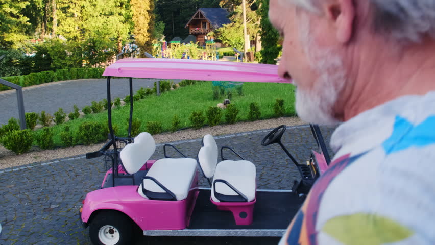 Senior man walks toward pink golf cart on cobblestone path near green hedge. Adult male stops beside small vehicle looking at seats and steering