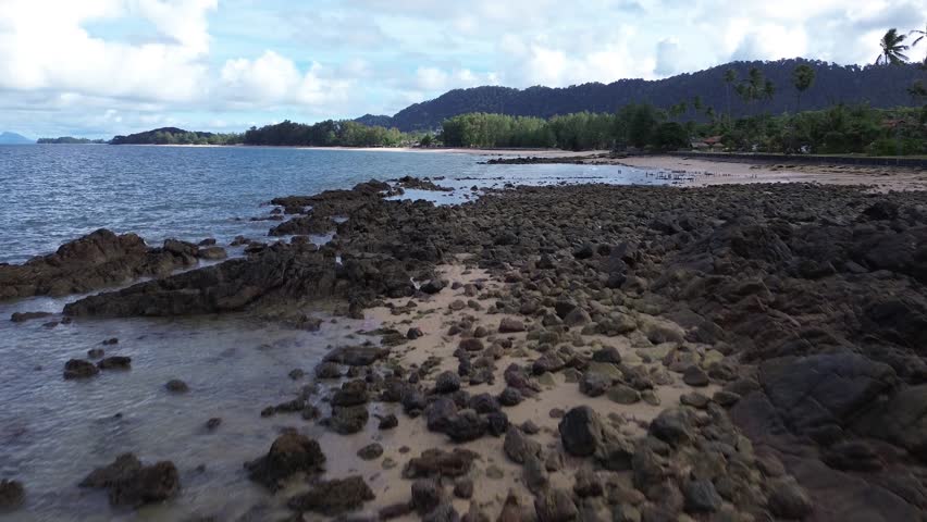 Aerial drone footage of a rocky shoreline at Long Beach Koh Lanta, Thailand, featuring lush palm trees in the foreground and tropical bungalows along the coast, with turquoise waters on a cloudy day.
