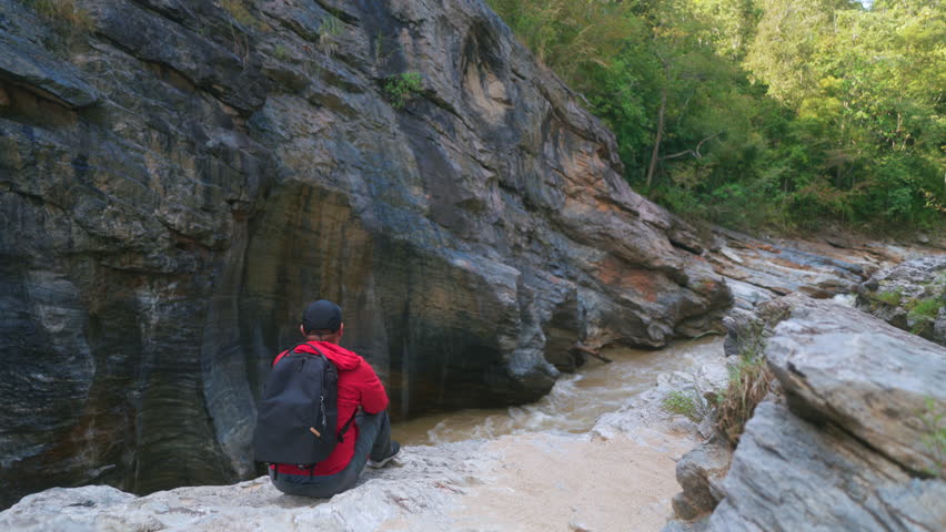 Happy asian male hiker stand on a light colored rock beside a gently flowing stream, holding a sturdy hiking stick inside the national park.