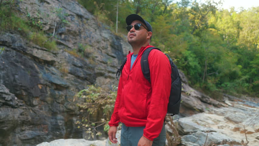 Happy asian male hiker stand on a light colored rock beside a gently flowing stream, holding a sturdy hiking stick inside the national park.