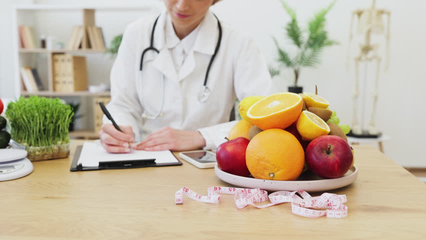A nutritionist is writing a diet plan with a plate of fresh fruits and a measuring tape on a wooden table.