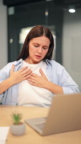 Woman experiences sharp chest pain and significant discomfort while working at her laptop. Clutches chest, displaying distress and concern, serious health issue requiring immediate attention.