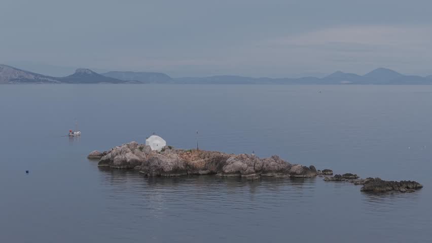 A morning drone capture showing a Hydra fisherman heading out to sea, passing a rocky islet with a small chapel in a calm, blue-toned scene.
