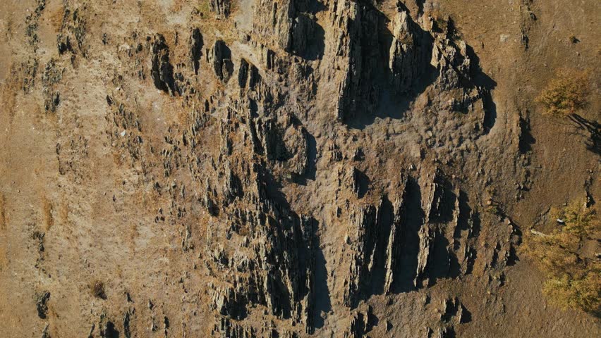 Măcin Mountains, Tulcea County, Romania - Jagged Rock Formations Rise from a Dry, Rugged Hillside, Casting Sharp Shadows Across the Textured Earth - Drone Ascend Shot