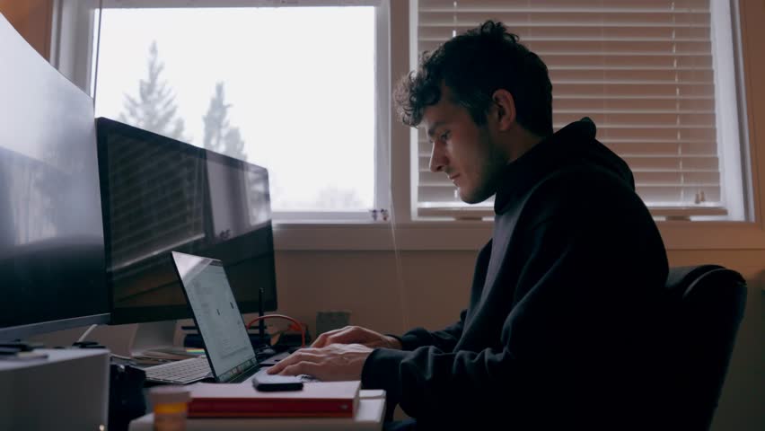 Static profile shot of a man working on a laptop in front of an external computer monitor at a home office desk with soft daylight