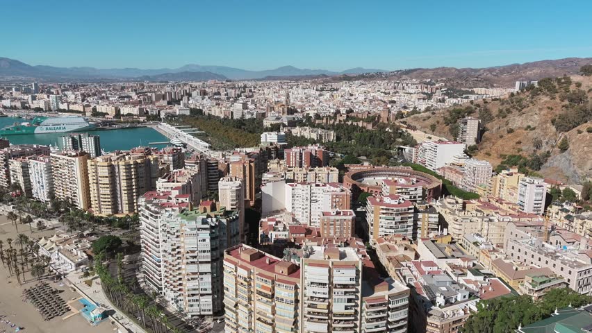 Aerial view captures the cityscape of Malaga Spain showcasing its buildings port and mountains under a clear blue sky on a sunny day