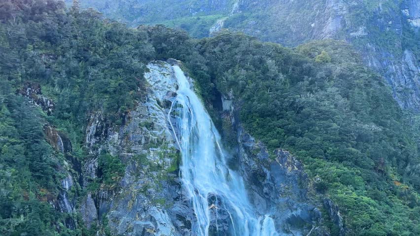 Sailing past a large waterfall in Milford Sound, Fiordland National Park, New Zealand. Towering cliffs, cascading water, and misty surroundings create a dramatic and scenic fjord view.