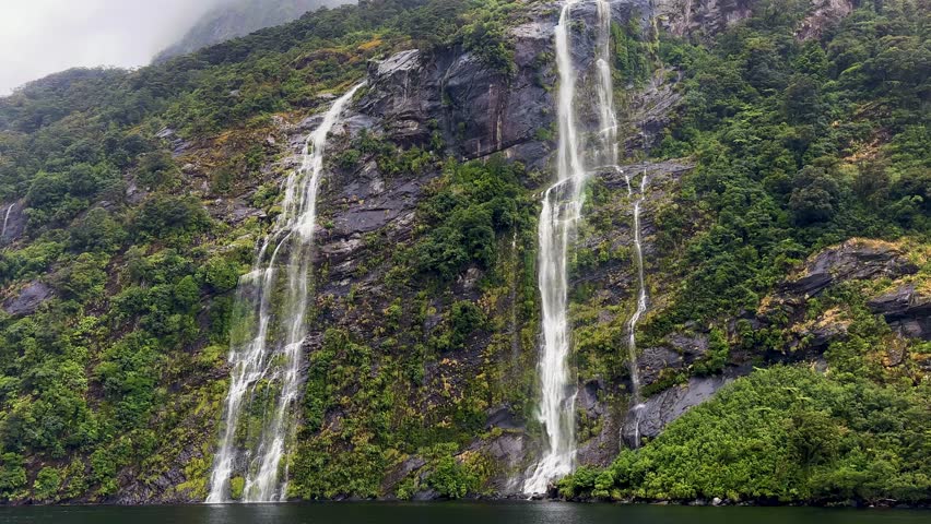 Looking up at towering twin waterfalls on a rainy day in Doubtful Sound, Fiordland National Park, New Zealand. Mist, rugged cliffs, and rushing water create a dramatic landscape.