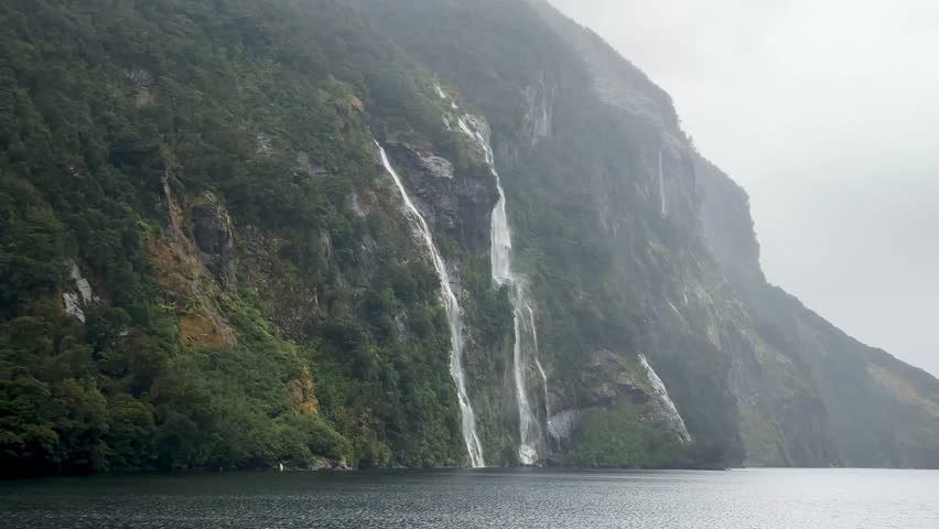 Twin waterfalls cascade down cliffs in Doubtful Sound, Fiordland National Park, New Zealand, on a moody, rainy day. Mist, rugged mountains, and rushing water create a dramatic scene.