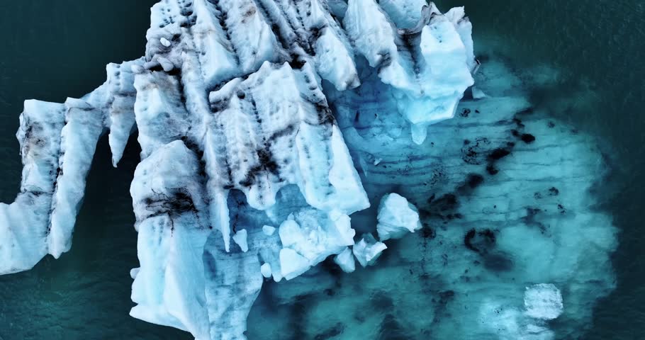 Top-down drone view of a fractured blue iceberg in Vatnajökull glacial lagoon, revealing deep crevasses and dark volcanic streaks beneath the cold water surface.