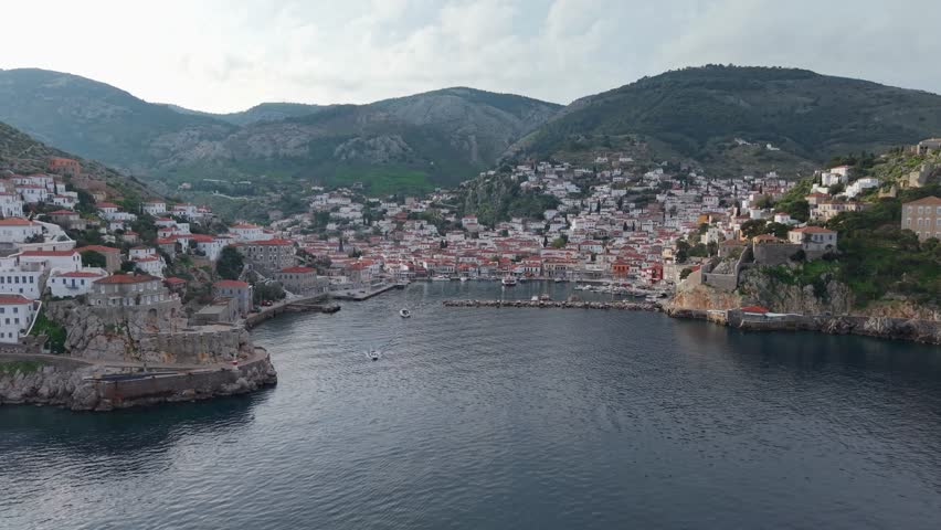 Morning drone orbit capturing daily life at Hydra port, with boats arriving and people moving along the harbor in soft early light.