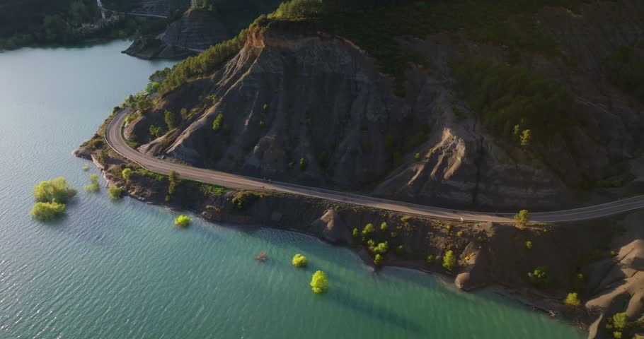 Aerial tracking shot of cyclists traversing the submerged ruins of the Antiguo Balneario de Tiermas and the arched masonry bridge of the A-1601 at the Yesa Reservoir, Spain.