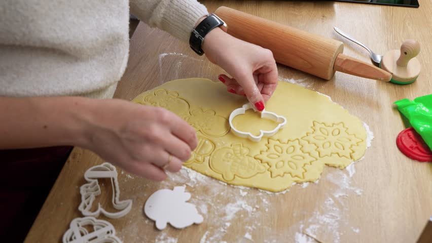 A close-up shot of hands pressing cookie cutters into dough, creating festive shapes. The scene captures warm natural lighting on a wooden table with baking tools nearby.