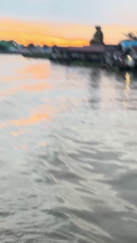 Wide tracking shot of a calm river at sunset with a silhouette of the city skyline and stilt houses on the bank. The video captures the orange sky reflecting on the rippling water surface