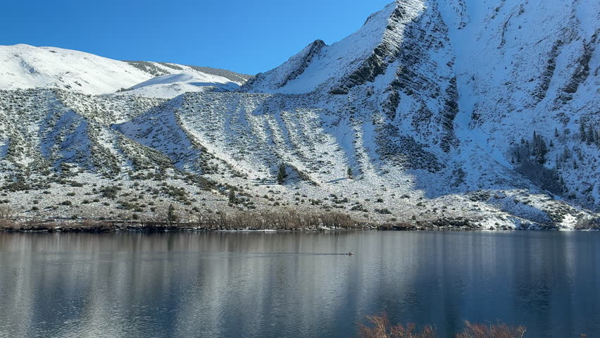 Convict Lake with a tiny kayak crossing mirror-like water below snow covered Eastern Sierra peaks on a bright, crisp winter day.