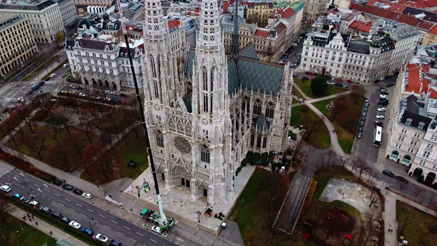 Aerial drone shot panning down to reveal Votivkirche cathedral in Vienna, highlighting its Gothic towers as surrounding city buildings, streets, and rooftops come into view under clear winter skies