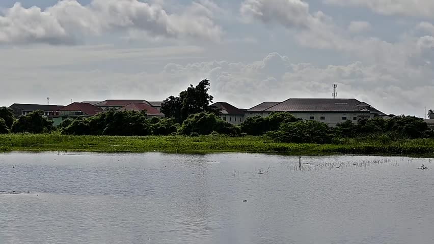 A tranquil body of water mirrors the landscape, featuring buildings nestled amongst lush trees and a sky filled with dramatic clouds.