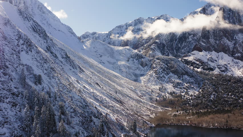 Snow-covered Sierra Nevada peaks rise above Convict Lake as a winter forest wraps the shoreline, captured in a crisp aerial flight along Highway 395 in California.