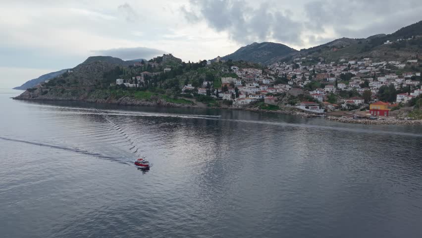 A winter morning drone view of Hydra, capturing the taxi boat used for daily transportation moving across the calm sea beside the island’s traditional harbor.
