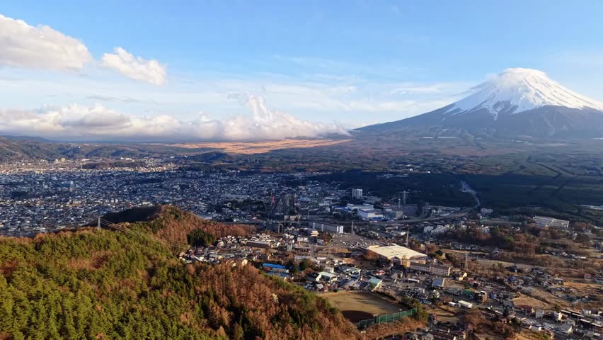 Aerial drone view of Fujikawaguchiko town, Japan, at the foothills of Mount Fuji in daylight, showcasing urban streets, greenery, and scenic mountains.