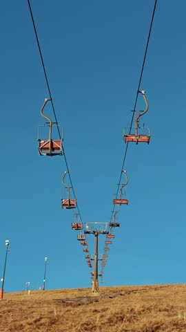 Orange lift chairs hanging on chairlift above dry slope under bright blue sky.