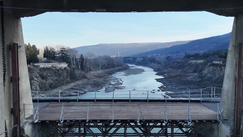 Aerial drone pullback revealing a hydroelectric dam with concrete gates and flowing river below. The camera moves backward to show the full industrial structure, surrounding water, and landscape. Hydropower infrastructure, renewable energy production, and water management system captured from above. Suitable for energy, sustainability, engineering, utilities, and environmental topics.