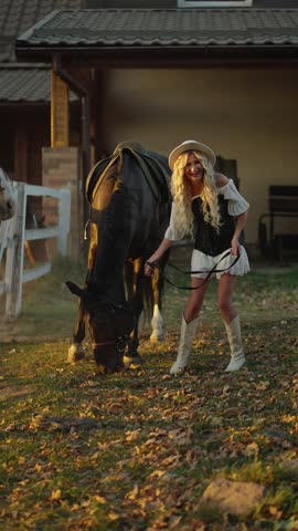 Smiling cowgirl leading a grazing horse near a countryside stable at golden hour, joyful rural lifestyle moment