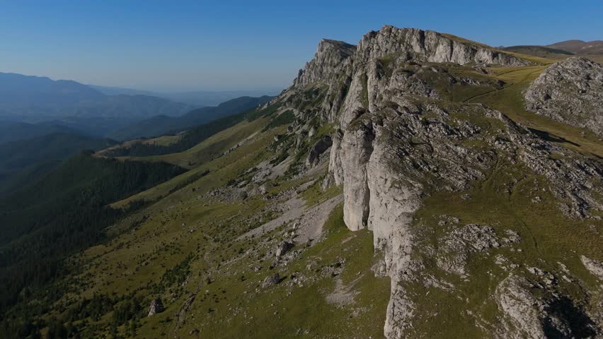 High-altitude aerial 4K footage capturing a rotation over the western ridge of the Bucegi Mountains in Romania. The shot highlights the spectacular geological formations of Strungile Mari and Coltii Tapului peaks, revealing numerous erosion towers and rugged rock faces. The late afternoon side light creates strong contrasts, dramatically accentuating the textures of the limestone cliffs and the depth of the alpine landscape.